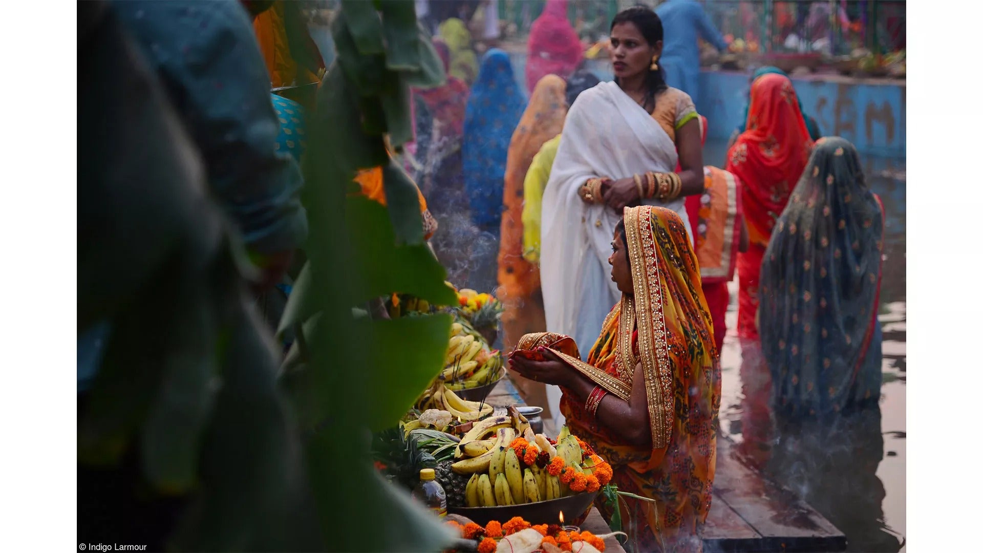 devotees stand in a body of water as the sun sets, giving offerings of food and prayer
