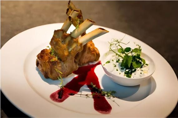Rack of lamb served with berry sauce and a side of herb-infused yogurt, garnished with fresh greens and thyme.