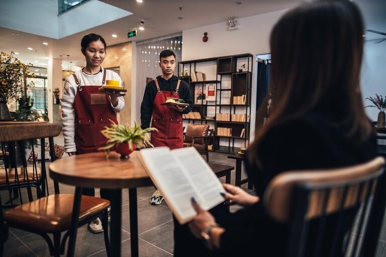 A woman reading a book in a restaurant whilst food is served.