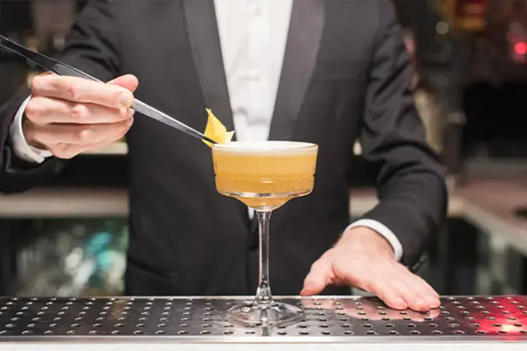 a hand garnishing a cocktail with tongs holding a piece of lemon zest