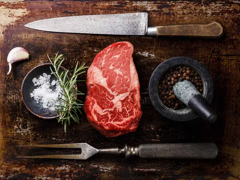 Ribeye steak on a board with seasoning, herbs, and an knife and fork.