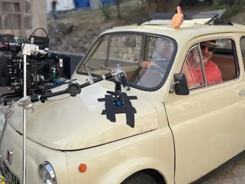 A man in a vintage white car with a video camera on the bonnet.